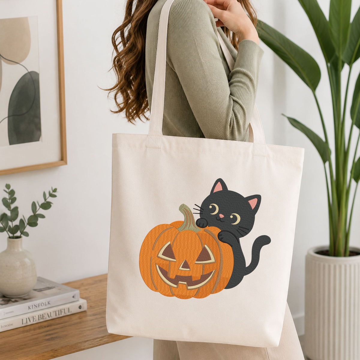 Woman carrying a white natural canvas tote bag featuring a cute black cat peeking behind a jack-o-lantern pumpkin machine embroidery design in a bright, modern interior setting