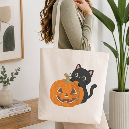 Woman carrying a white natural canvas tote bag featuring a cute black cat peeking behind a jack-o-lantern pumpkin machine embroidery design in a bright, modern interior setting