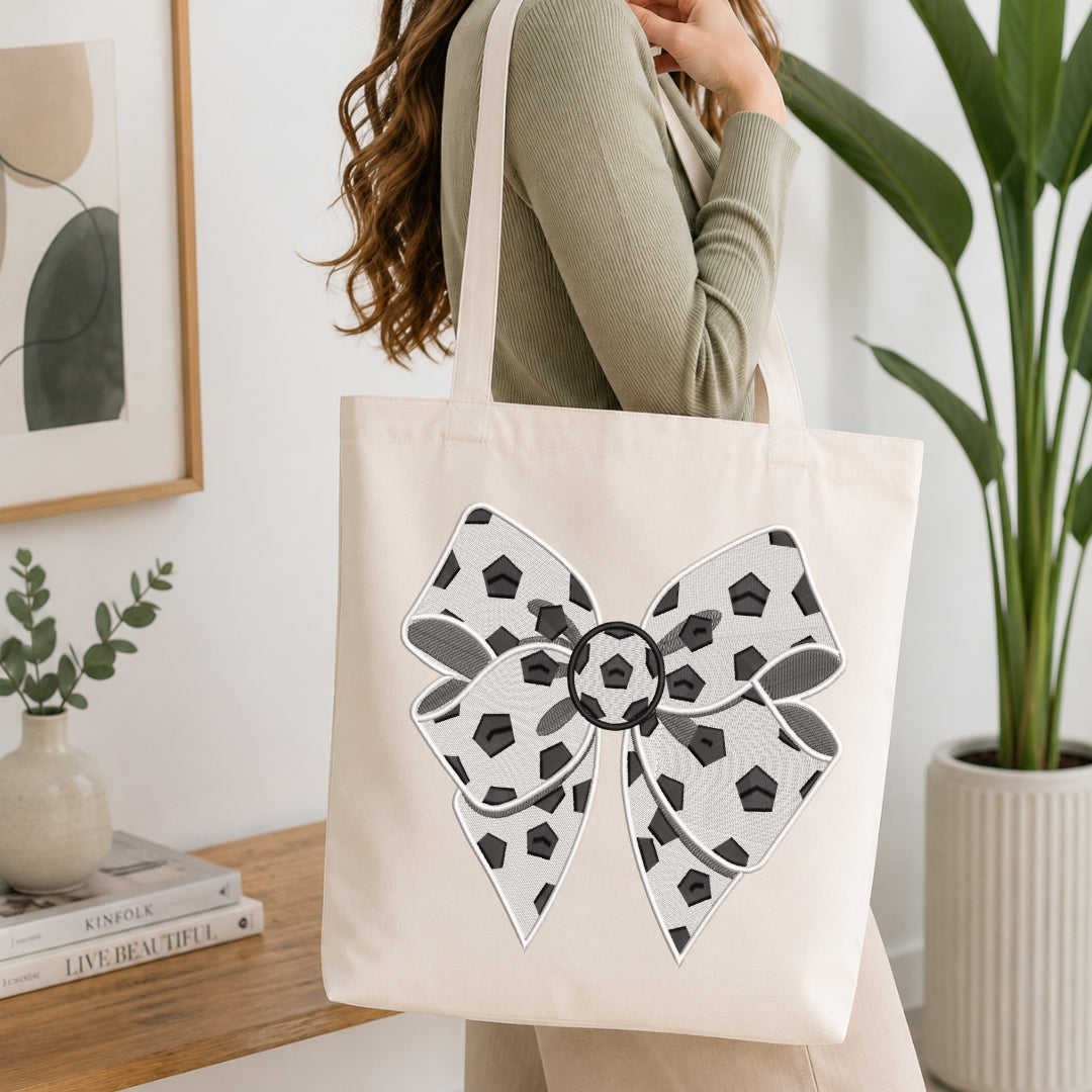 A woman wearing a sage green ribbed long-sleeve top carries a cream canvas tote bag with a large black and white patterned bow graphic on the front, set in a bright, modern room with a wooden table and potted plants.