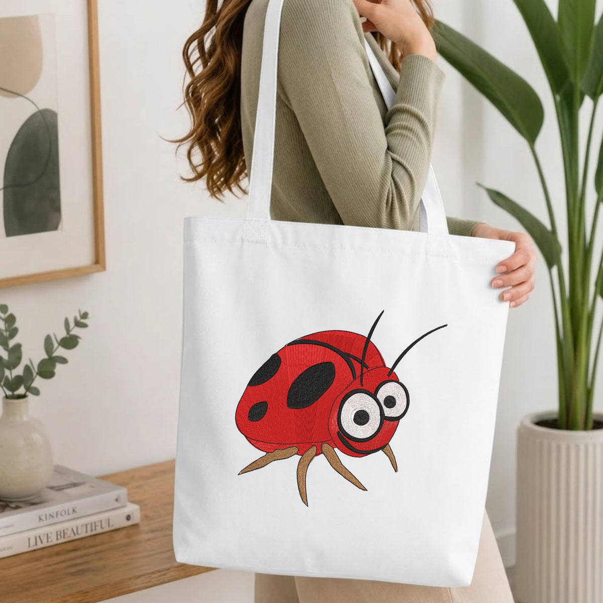 Woman carrying a white natural canvas tote bag featuring an embroidered cute ladybug design in a bright, modern interior setting
