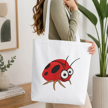 Woman carrying a white natural canvas tote bag featuring an embroidered cute ladybug design in a bright, modern interior setting