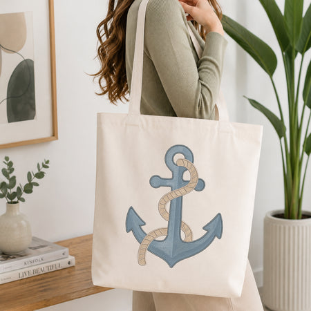 A woman wearing a sage green ribbed sweater carries a cream canvas tote bag with a large embroidered blue anchor and beige rope design on the front, set in a bright, modern room with a wooden table, framed art, and a potted plant.