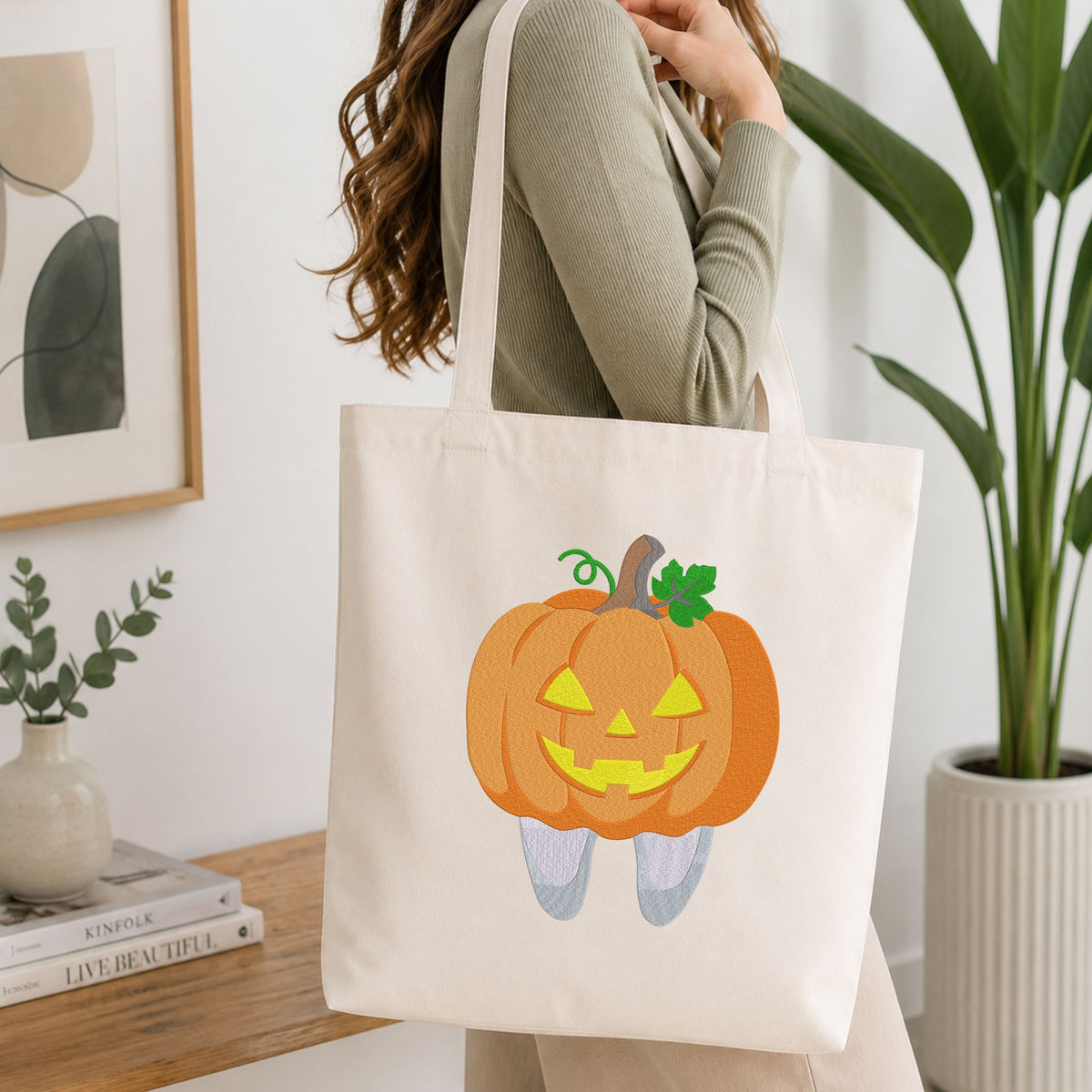 Woman carrying a white natural canvas tote bag featuring a cute jack-o-lantern pumpkin tooth Halloween dental machine embroidery design in a bright, modern interior setting   