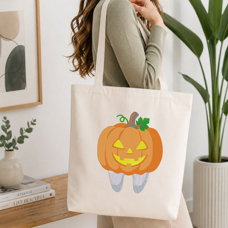 Woman carrying a white natural canvas tote bag featuring a cute jack-o-lantern pumpkin tooth Halloween dental machine embroidery design in a bright, modern interior setting   