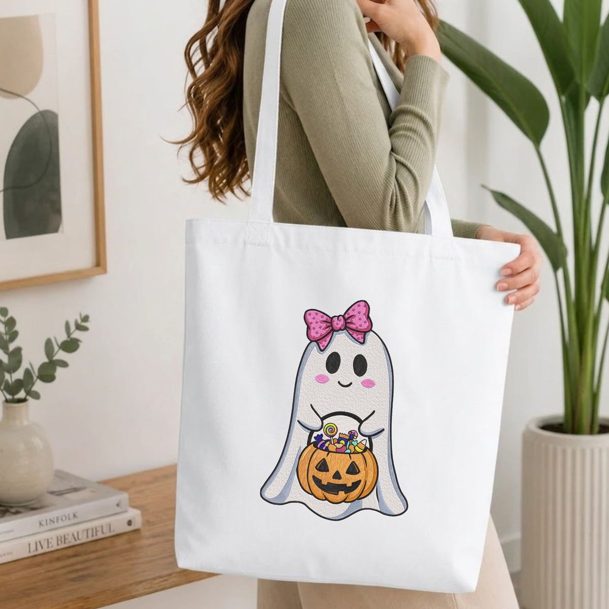 Woman carrying a white natural canvas tote bag featuring a cute ghost machine embroidery design in a bright, modern interior setting