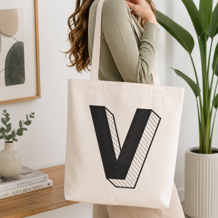 A woman wearing a sage green ribbed long-sleeve top carries a cream canvas tote bag with a large black block letter 'V' printed on the front, set in a bright, modern room with a wooden console table, framed art, and a potted plant.