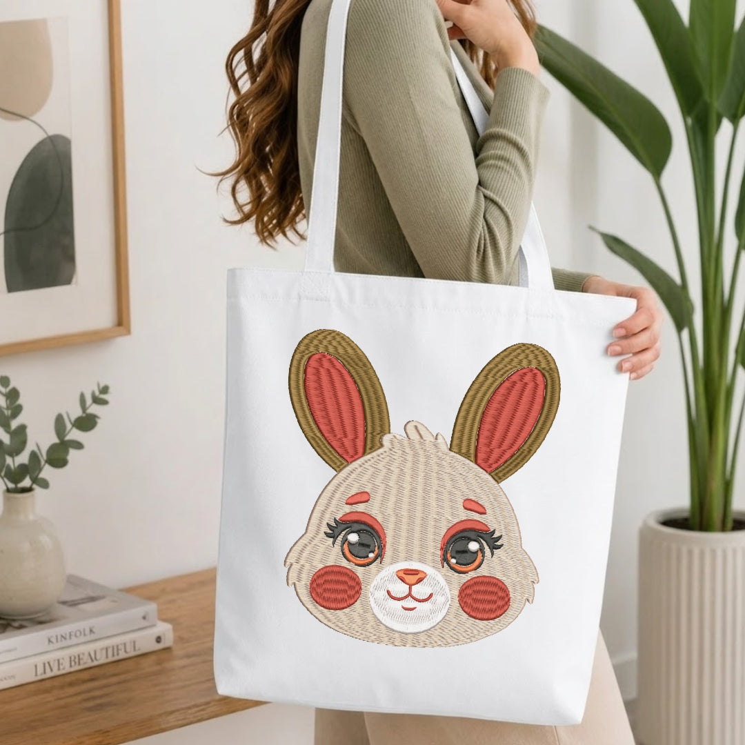 A woman holds a white canvas tote bag featuring a large embroidered bunny face with pink inner ears and cheeks. The scene is set in a bright, modern room with a wooden table, books, and a potted plant in the background.