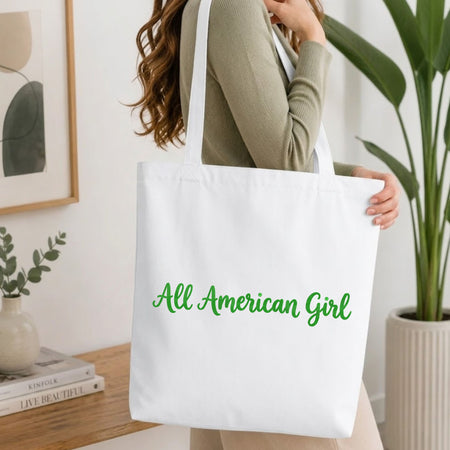 Woman carrying a white natural canvas tote bag featuring an embroidered green "All American Girl" text design in a bright, modern interior setting.