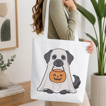 A woman holds a white canvas tote bag featuring a cartoon graphic of a pug dressed in a ghost costume and holding a jack-o'-lantern pumpkin. The scene is set in a bright, modern room with a wooden table, books, and a potted plant.
