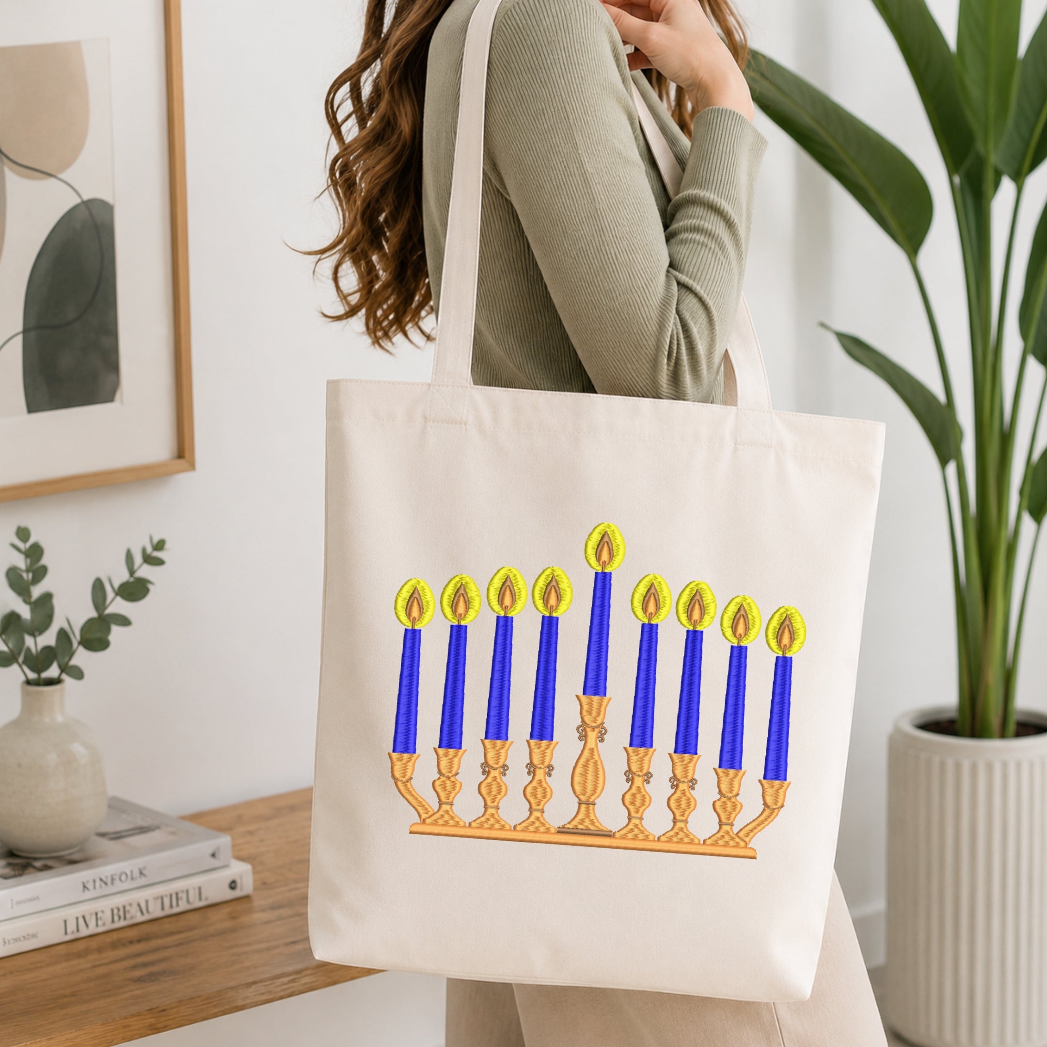 A woman holds a cream canvas tote bag featuring a printed Hanukkah menorah with blue candles and gold candlesticks. The scene is set in a bright, modern room with a wooden table, books, a framed print, and a potted plant.