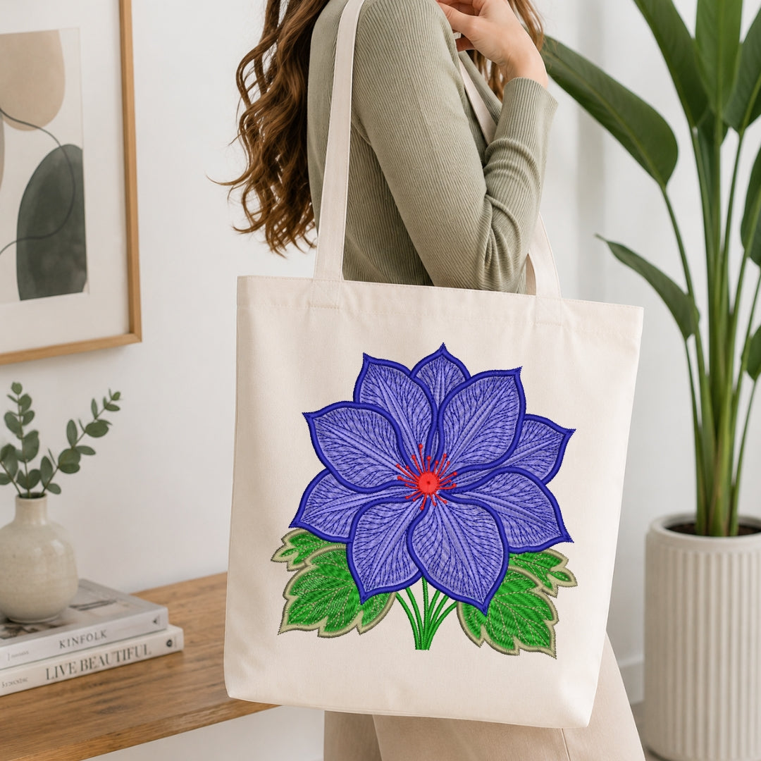 A woman wearing a sage green ribbed sweater carries a cream canvas tote bag with a large embroidered blue lotus flower design in purple, green, and red, set against a bright, modern interior background with a wooden table and potted plants.