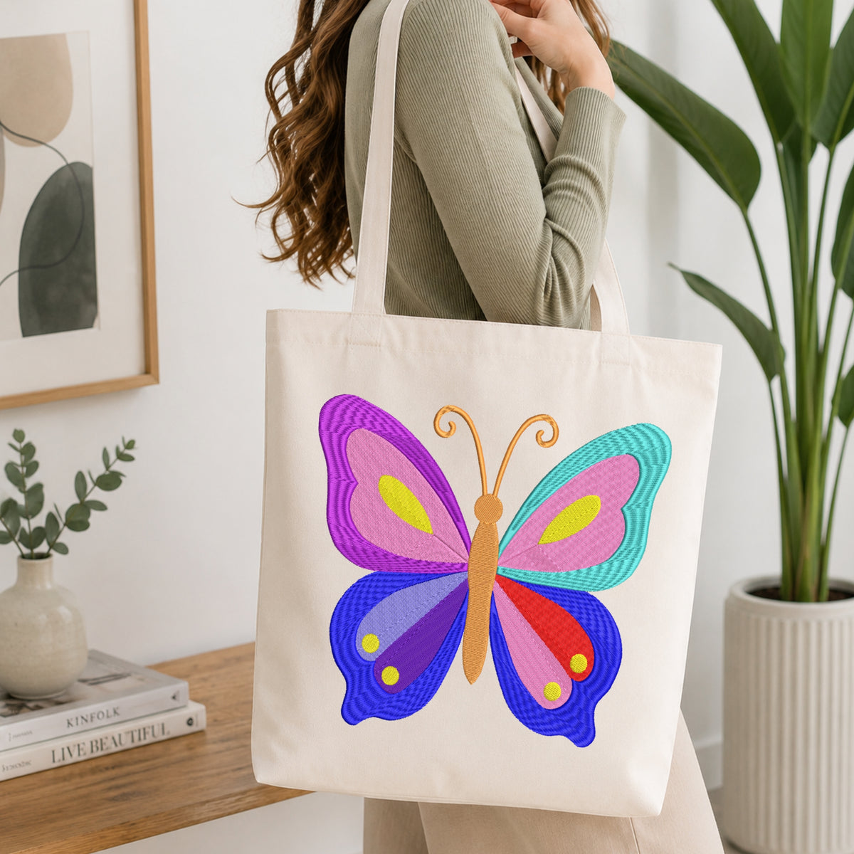 A woman holds a cream canvas tote bag featuring a colorful, textured butterfly design in purple, pink, blue, red, and yellow. The scene is set in a bright, modern room with a wooden table, books, a potted plant, and framed wall art.