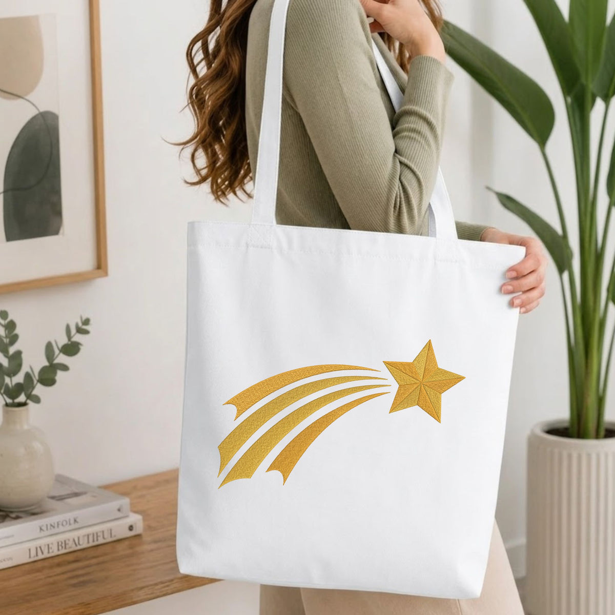 Woman carrying a white natural canvas tote bag featuring an embroidered golden shooting star design in a bright, modern interior setting