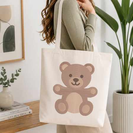 A woman wearing a sage green ribbed long-sleeve top carries a cream canvas tote bag featuring a cute brown teddy bear graphic. The scene is set in a bright, modern room with a wooden table, a small potted plant, and a large green houseplant in the background.
