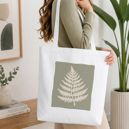 Woman carrying a white natural canvas tote bag featuring an embroidered sage green fern leaf design in a bright, modern interior setting.