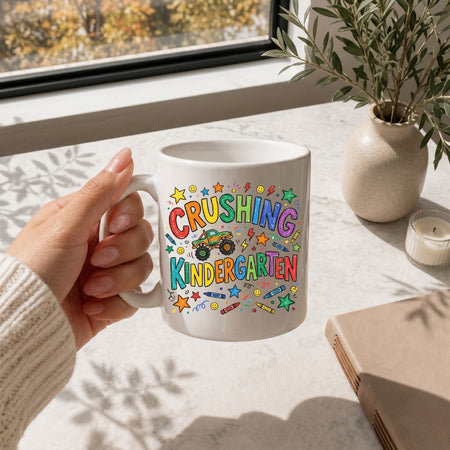 A hand holds a white ceramic mug with a colorful 'CRUSHING KINDERGARTEN' graphic featuring a monster truck and stars, set on a sunlit windowsill with a plant and candle in the background.