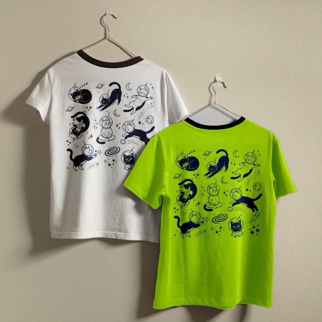 Two short-sleeve ringer T-shirts hanging on hangers against a plain beige wall. The back of one shirt is white with a dark brown collar and a black cat-themed graphic print, and the other is bright neon yellow-green with a black collar and the same black cat-themed graphic print.