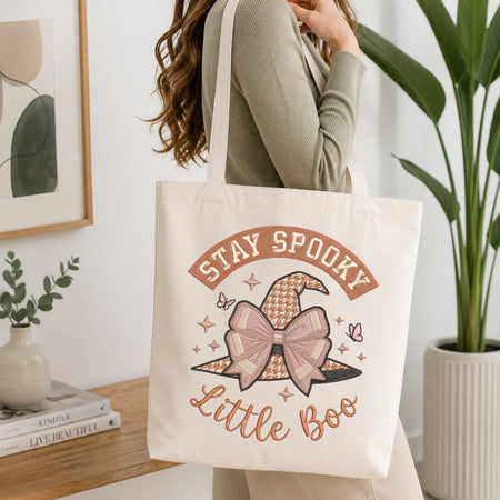 A woman wearing a sage green ribbed sweater carries a cream canvas tote bag with a 'Stay Spooky Little Boo' Halloween graphic featuring a patterned witch hat and bow.