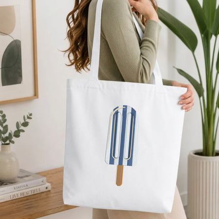 Woman carrying a white natural canvas tote bag featuring an embroidered blue and white striped ice cream popsicle design in a bright, modern interior setting.