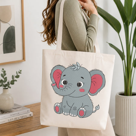 A person wearing a ribbed sage green top carries a cream canvas tote bag featuring a cute cartoon gray baby elephant with pink ears and pink feet. The scene is set in a bright, modern room with a wooden table, books, a framed print, and a potted plant.