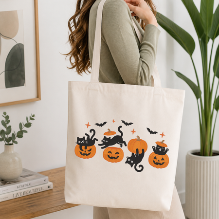 Woman carrying a white natural canvas tote bag featuring a playful black cats and pumpkins machine embroidery design in a bright, modern interior setting