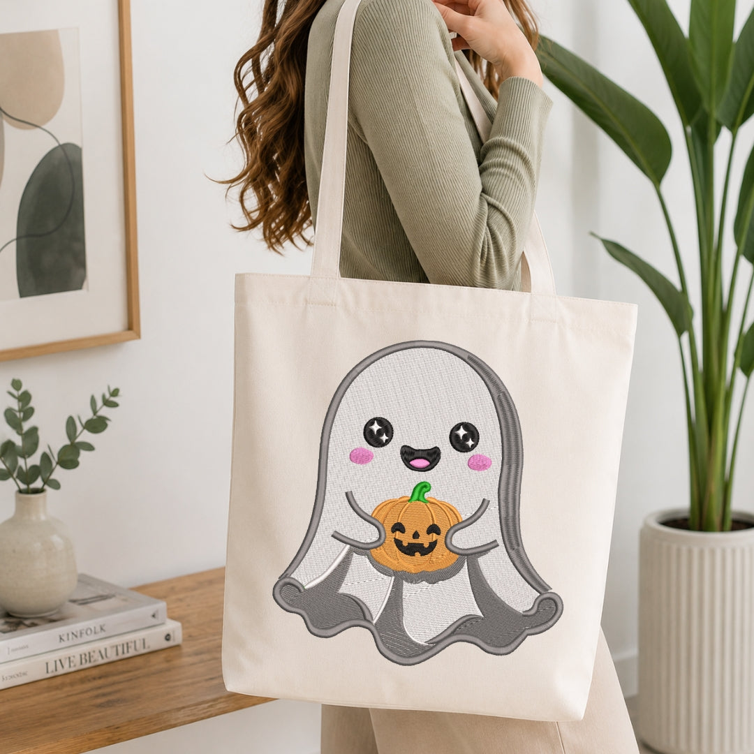 A person wearing a sage green ribbed long-sleeve top carries a cream canvas tote bag with a cute embroidered ghost holding a jack-o'-lantern design. The scene is set in a bright, modern room with a wooden table, books, and a potted plant.