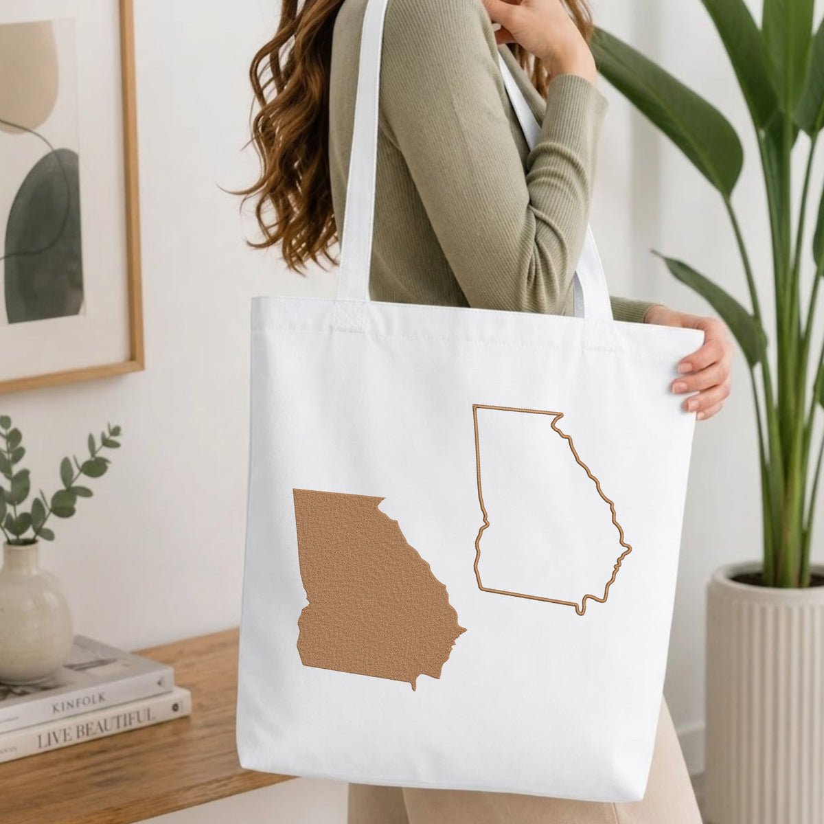 A woman wearing a sage green ribbed sweater carries a white canvas tote bag featuring a textured brown Georgia state silhouette and a brown outline of the same state on the front, set in a bright, modern room with a wooden table, books, and a potted plant.
