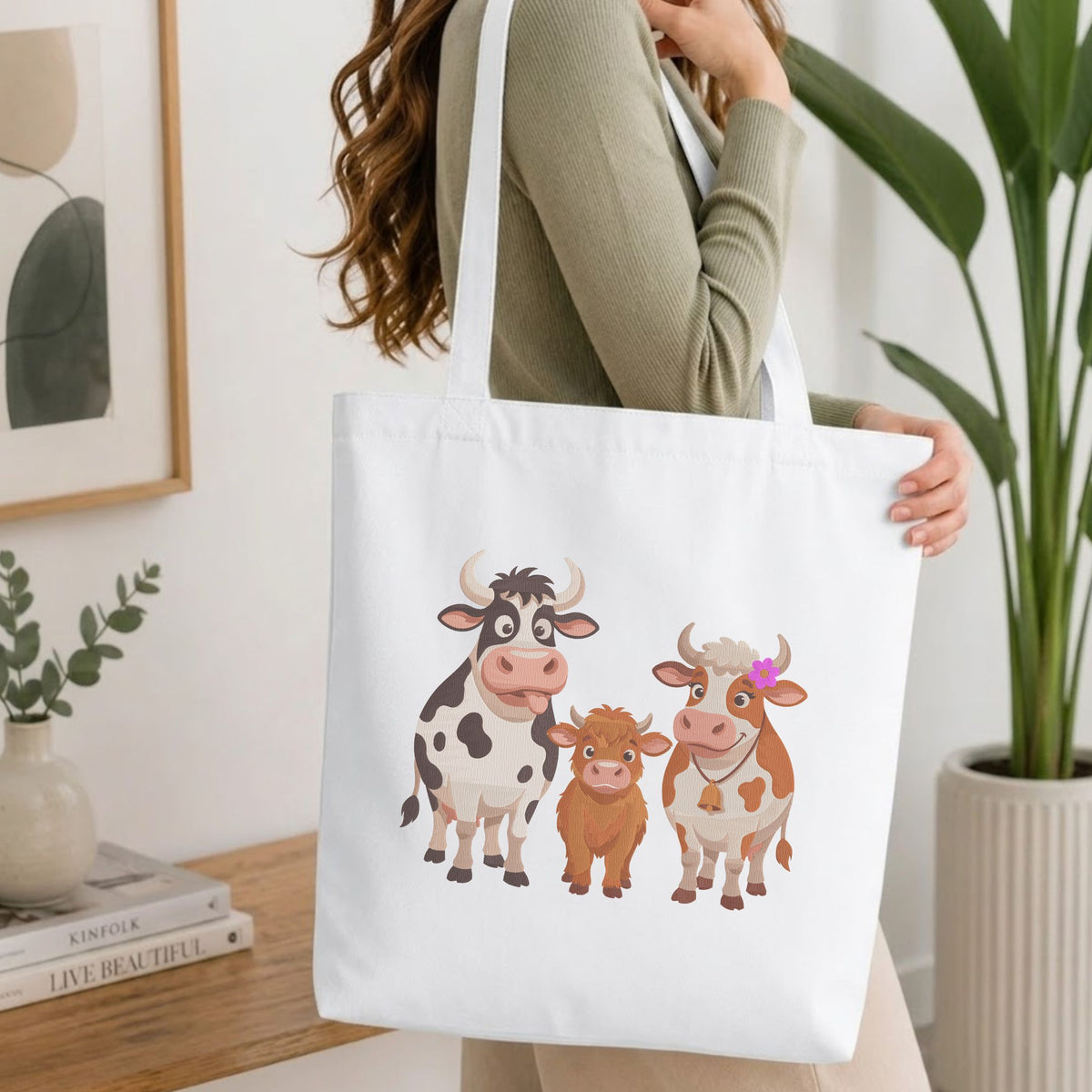 Woman carrying a white natural canvas tote bag featuring a cute cow family machine embroidery design in a bright, modern interior setting