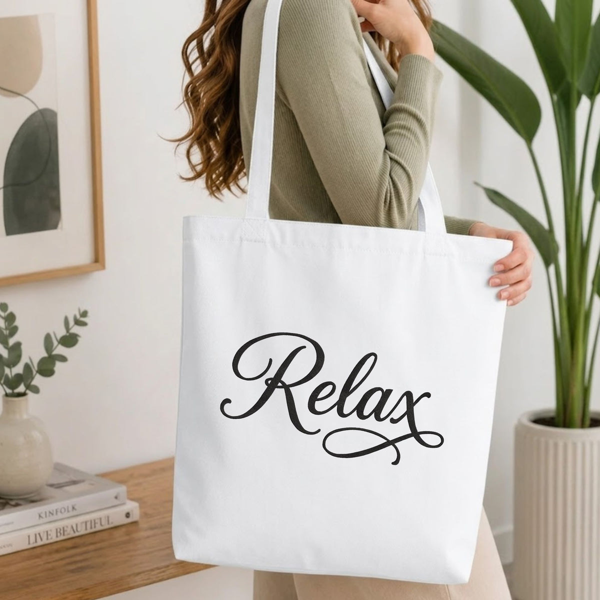 Woman carrying a white natural canvas tote bag featuring an embroidered "Relax" script design in a bright, modern interior setting   