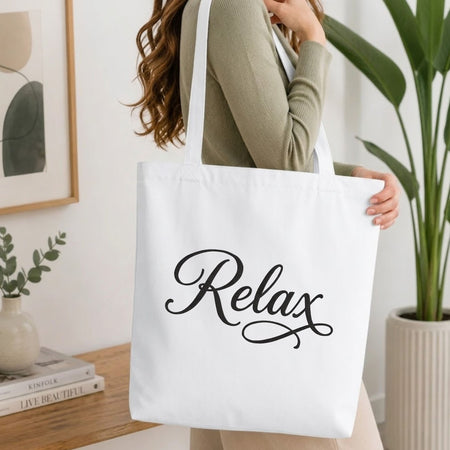 Woman carrying a white natural canvas tote bag featuring an embroidered "Relax" script design in a bright, modern interior setting   