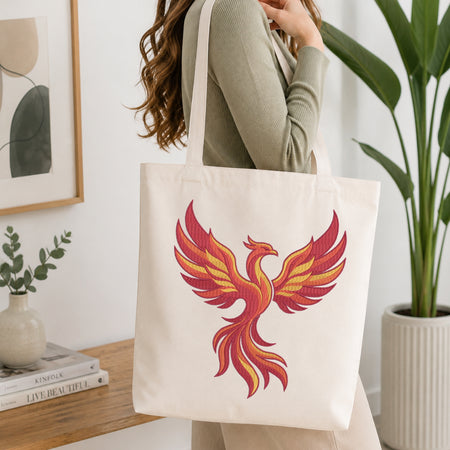 An off-white canvas tote bag with a large embroidered phoenix design in red, orange, and yellow, carried by a woman in a ribbed sage green top in a bright, modern room.