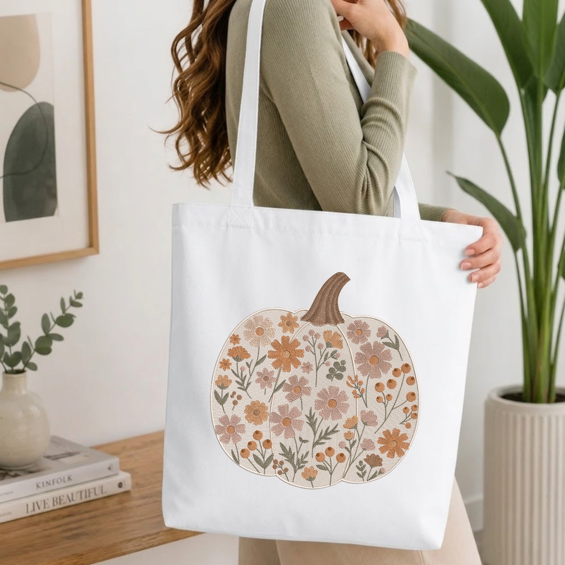 A woman in a sage green ribbed sweater carries a white canvas tote bag decorated with a large floral pumpkin design in muted orange, beige, and brown tones. The background is a bright, modern room with a wooden table, books, a potted plant, and framed wall art.
