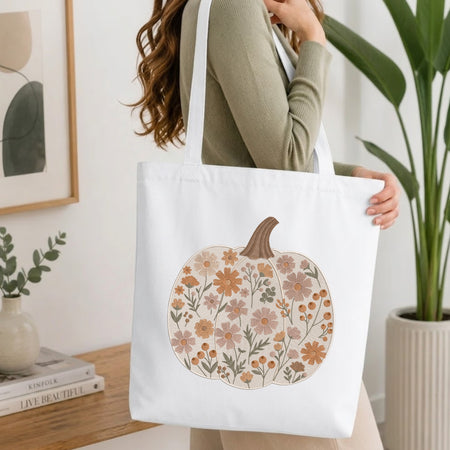 A woman in a sage green ribbed sweater carries a white canvas tote bag decorated with a large floral pumpkin design in muted orange, beige, and brown tones. The background is a bright, modern room with a wooden table, books, a potted plant, and framed wall art.