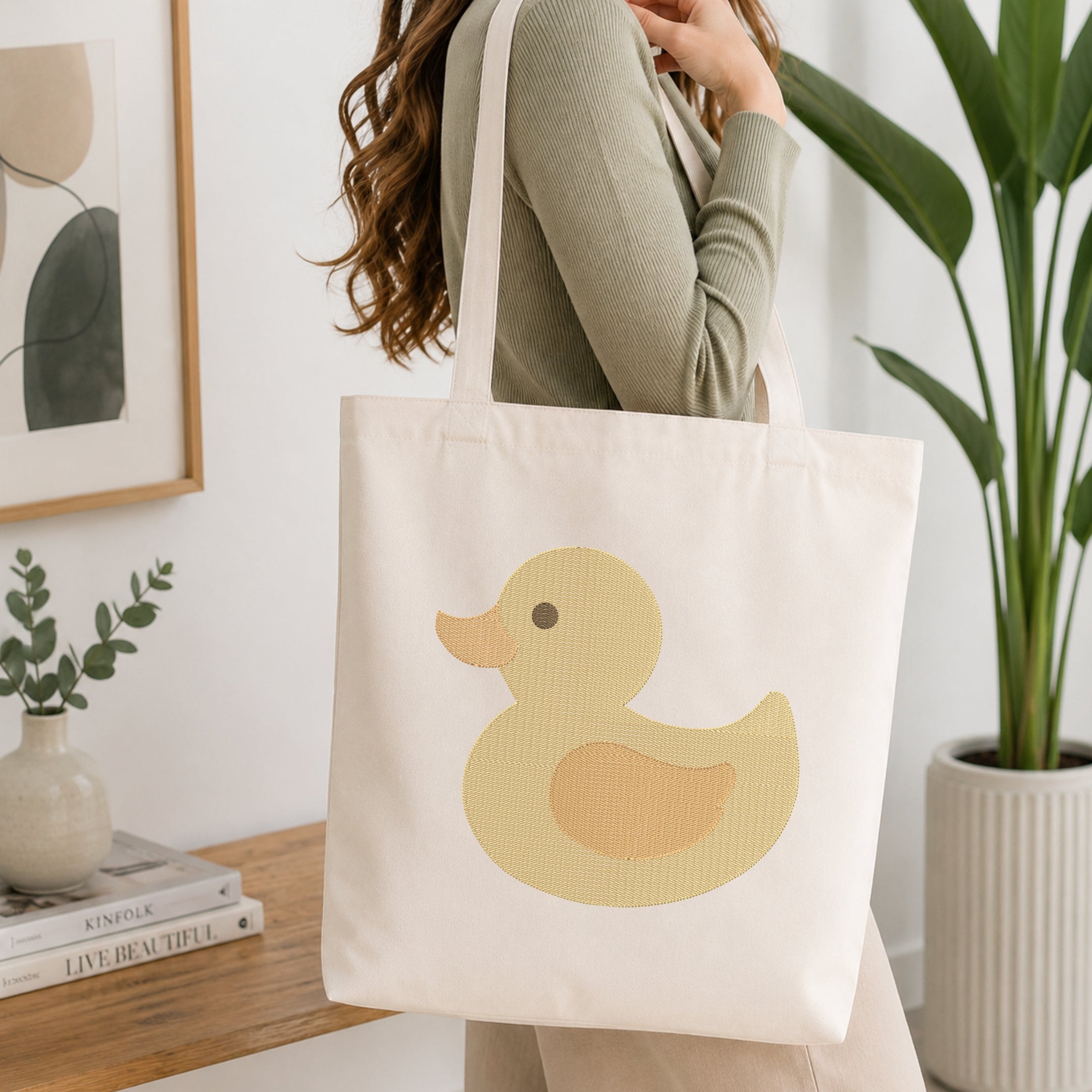 A woman wearing a sage green ribbed top carries a cream canvas tote bag with a large yellow duck graphic on the front, set in a bright, modern room with a wooden table and potted plants.