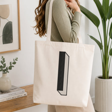 A woman wearing a sage green ribbed long-sleeve top carries a large off-white canvas tote bag with a black geometric block graphic on the front, set in a bright, modern room with a wooden console table, framed art, and a potted plant.