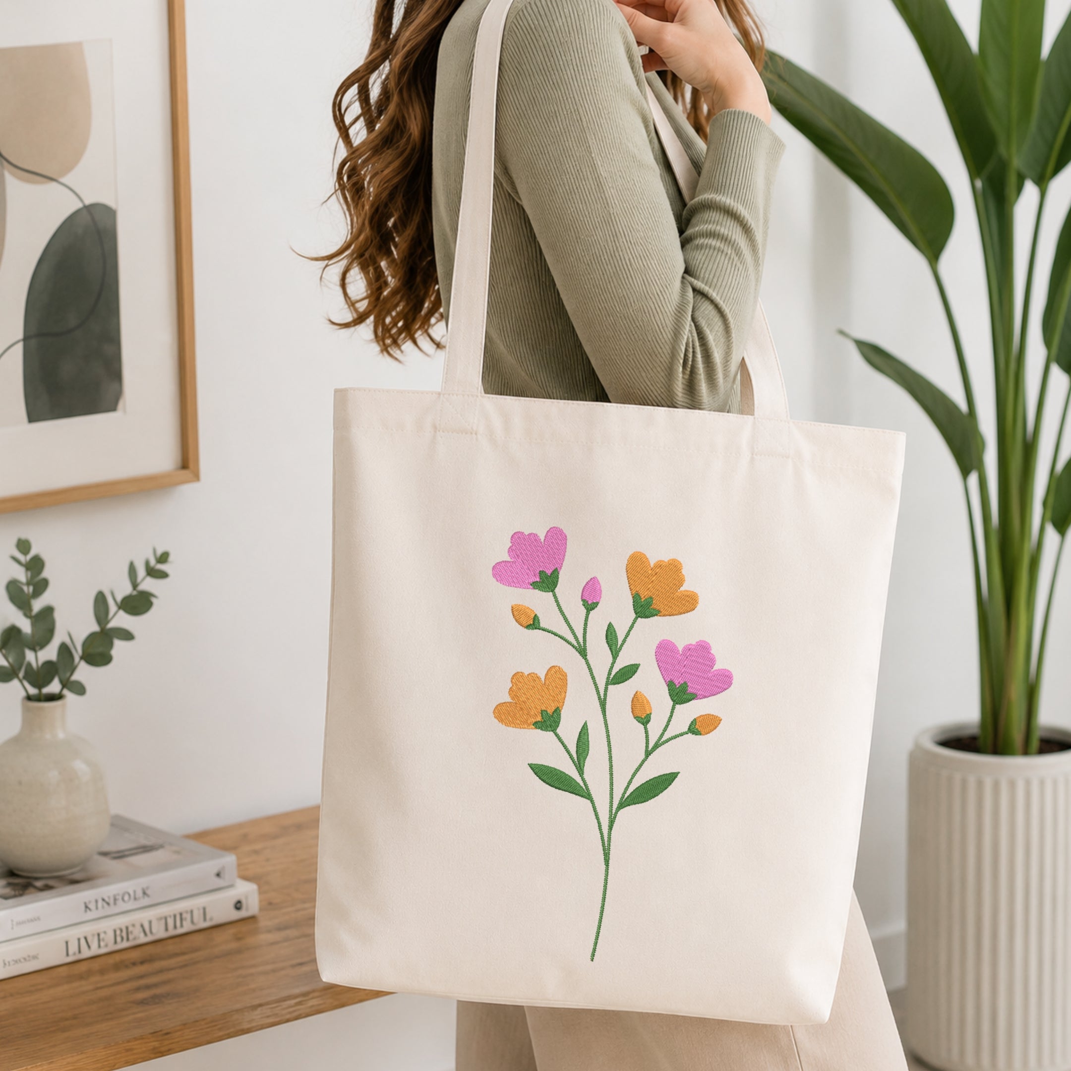 A woman wearing a sage green ribbed sweater carries a cream canvas tote bag with a colorful floral print of pink, orange, and yellow flowers with green leaves, set in a bright, modern room with a wooden table and potted plants.