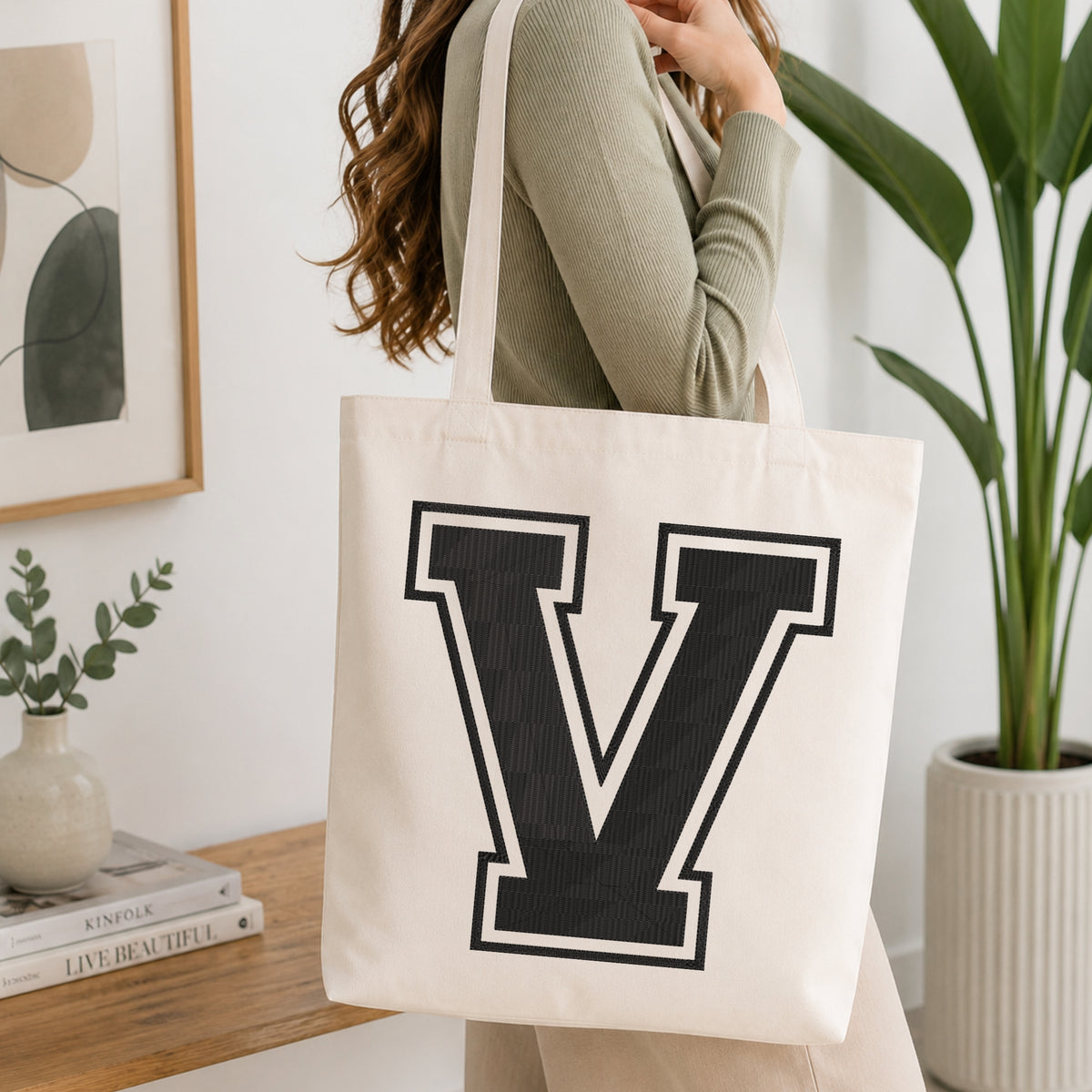 A woman wearing a sage green ribbed long-sleeve top carries a large off-white canvas tote bag with a bold black block letter 'V' outlined in white on the front, set in a bright, modern room with a wooden console table, framed art, and a potted plant.