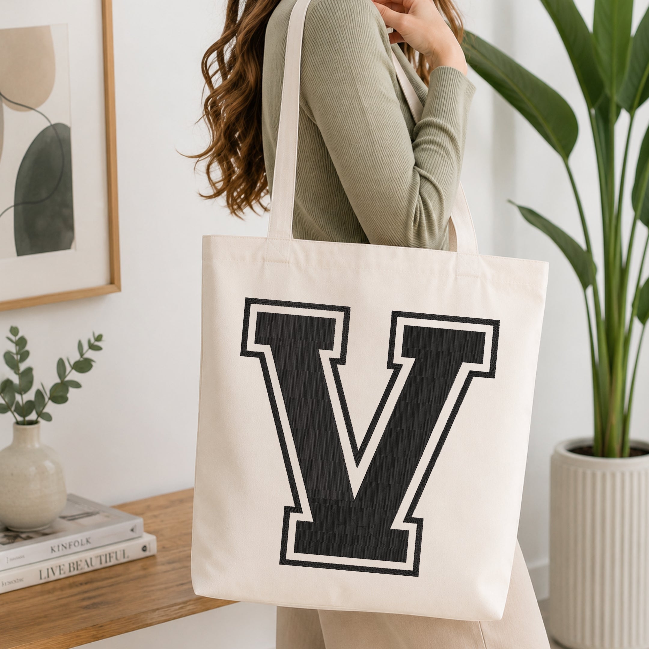A woman wearing a sage green ribbed long-sleeve top carries a large off-white canvas tote bag with a bold black block letter 'V' outlined in white on the front, set in a bright, modern room with a wooden console table, framed art, and a potted plant.