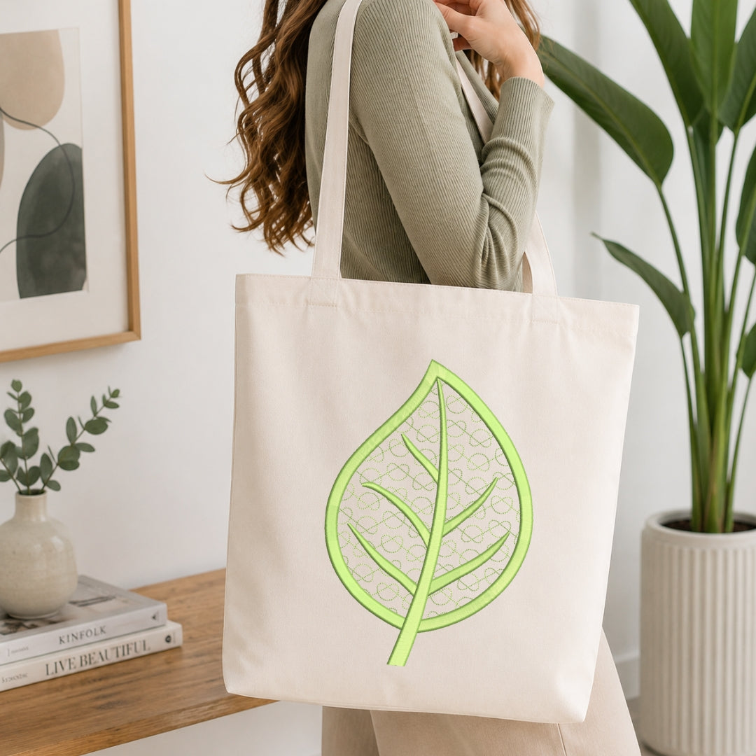 A woman wearing a sage green ribbed sweater carries a cream canvas tote bag with a bright green leaf design printed on the front, set in a bright, modern room with a wooden table and potted plants.