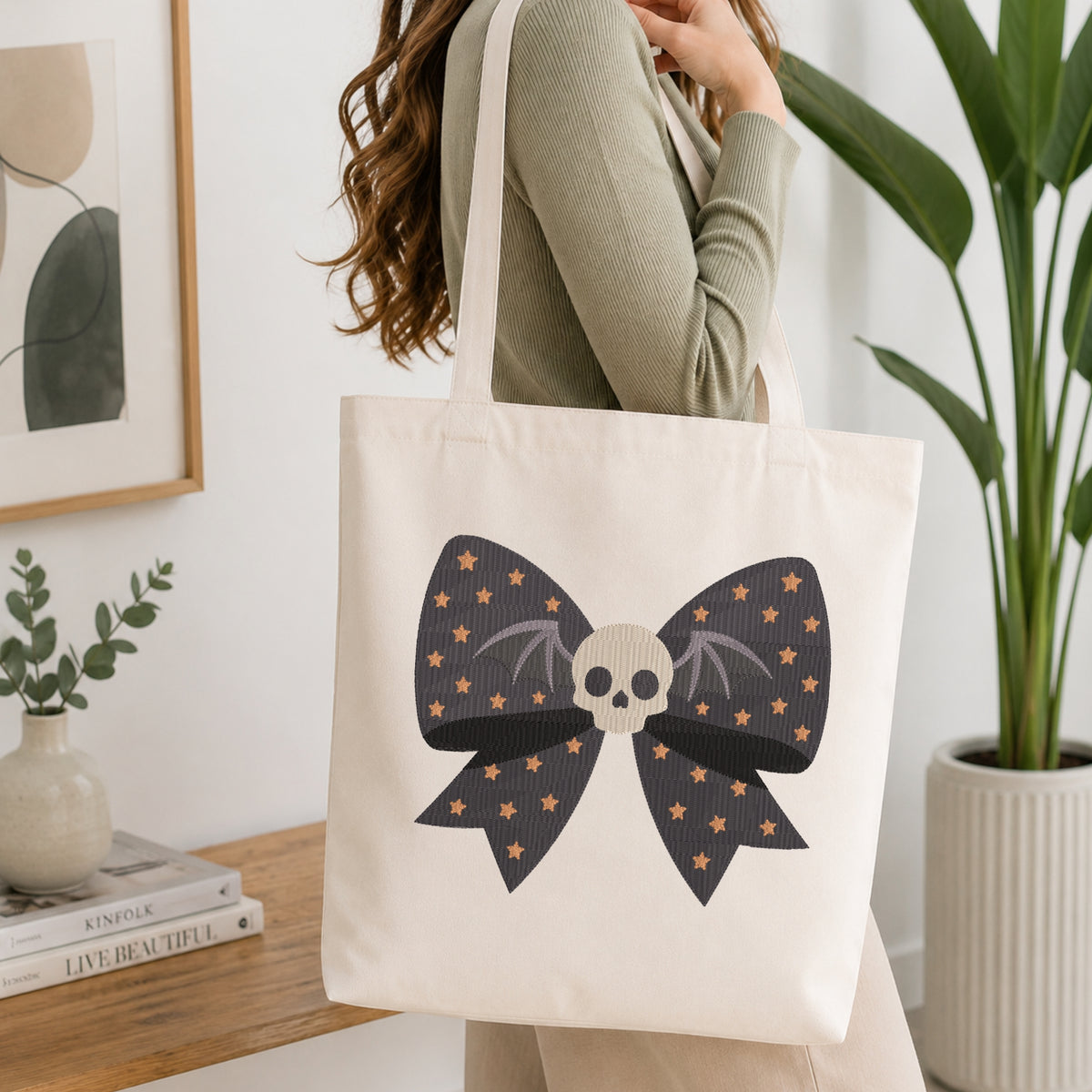 A person wearing a green long-sleeve top and carrying a natural canvas tote bag printed with the gothic starry skull bow machine embroidery graphic.