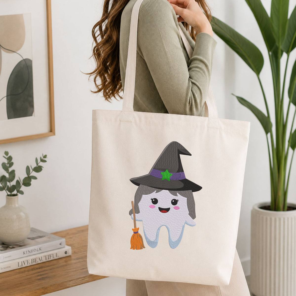 Woman carrying a white natural canvas tote bag featuring a cute witch tooth Halloween dental machine embroidery design in a bright, modern interior setting