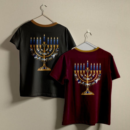 Two short-sleeve t-shirts hanging on hangers against a plain beige wall, showing the back designs. One shirt is black with a gold-trimmed collar and features a large menorah graphic with blue candles and white floral details.