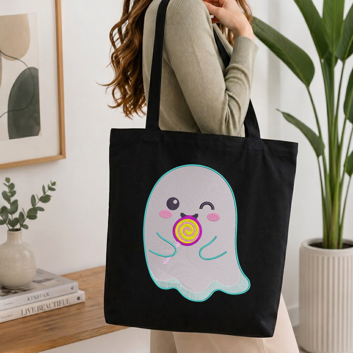 A person wearing a beige ribbed long-sleeve top carries a black canvas tote bag with a cute cartoon ghost graphic holding a pink and yellow lollipop. The background shows a cozy indoor setting with a wooden table, books, a framed print, and a potted plant.