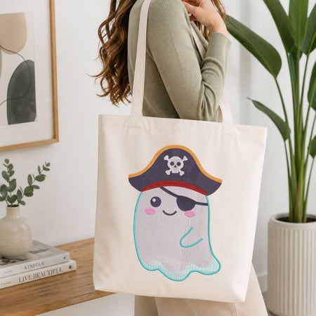 A woman holds a cream canvas tote bag featuring a cute embroidered pirate ghost character. The character has a dark blue pirate hat with a white skull and crossbones, a black eye patch, pink cheeks, and a light blue stitched outline.