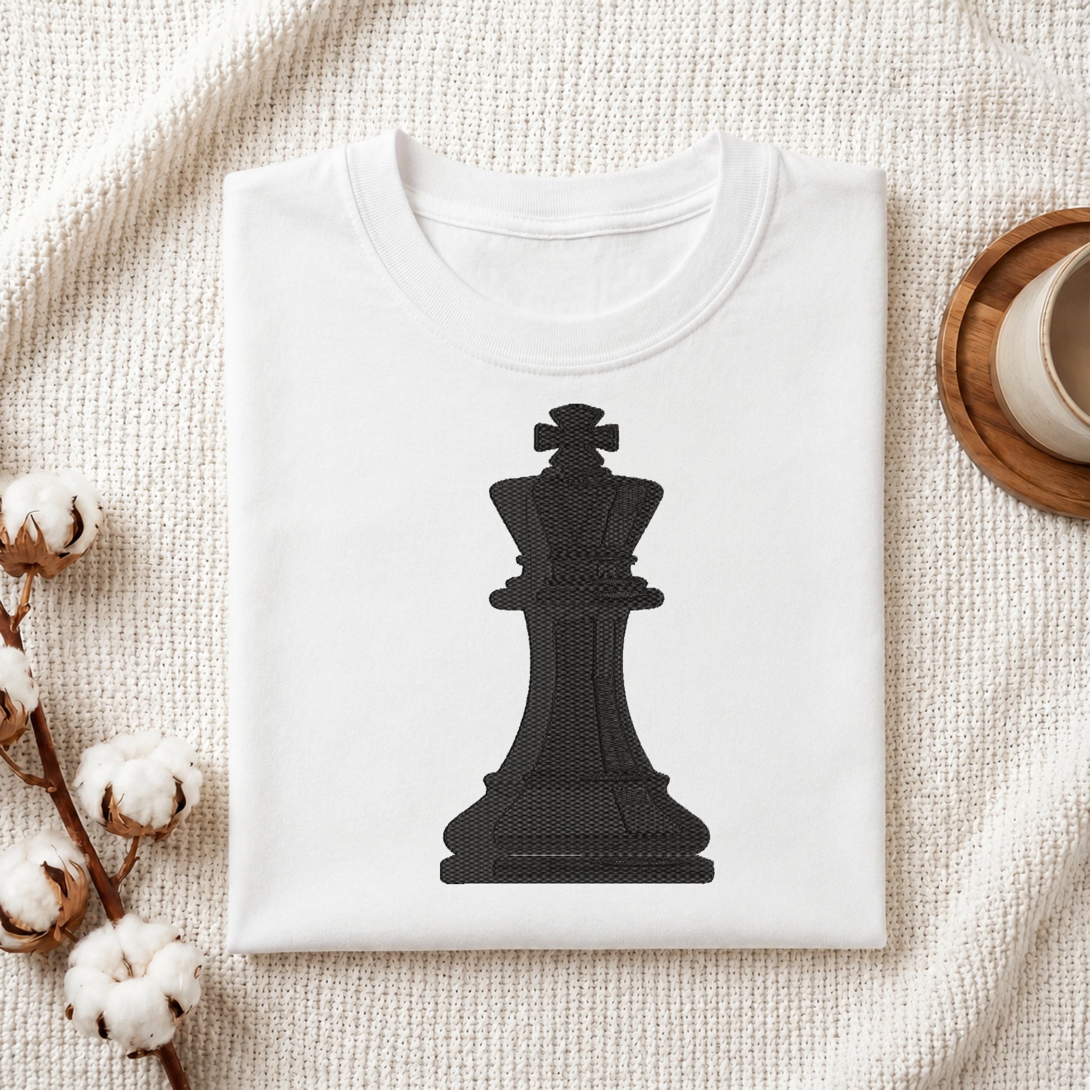 A folded white T-shirt with a large black chess king graphic on the front, styled on a textured cream fabric with cotton stems and a cup edge visible in the corners.