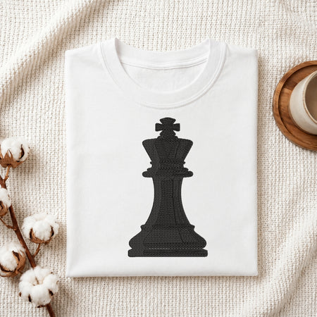 A folded white T-shirt with a large black chess king graphic on the front, styled on a textured cream fabric with cotton stems and a cup edge visible in the corners.