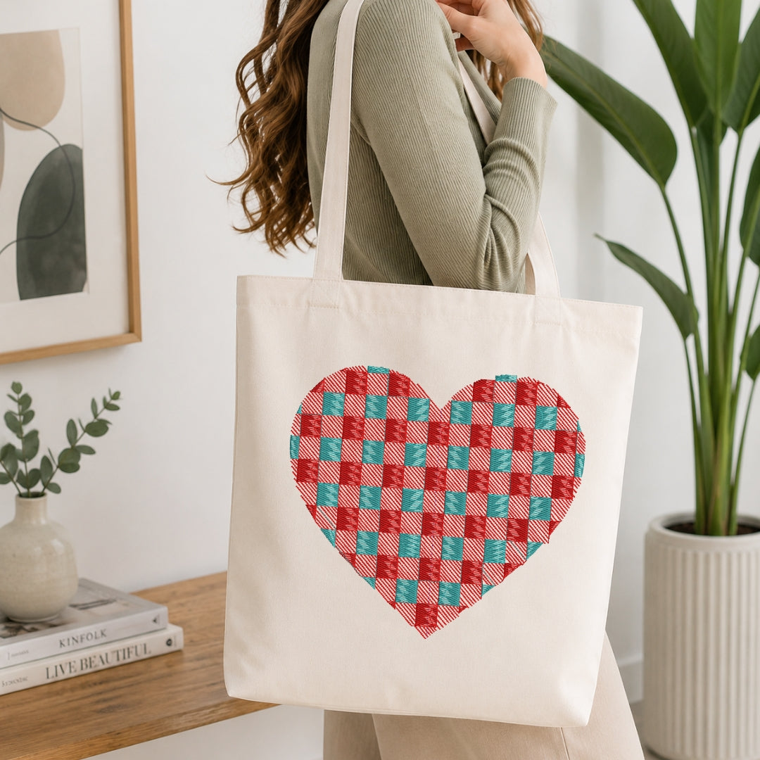 A woman wearing a sage green ribbed sweater carries a cream canvas tote bag with a large red and teal checkered heart design on the front, set in a bright, modern room with a wooden table, books, and a potted plant.