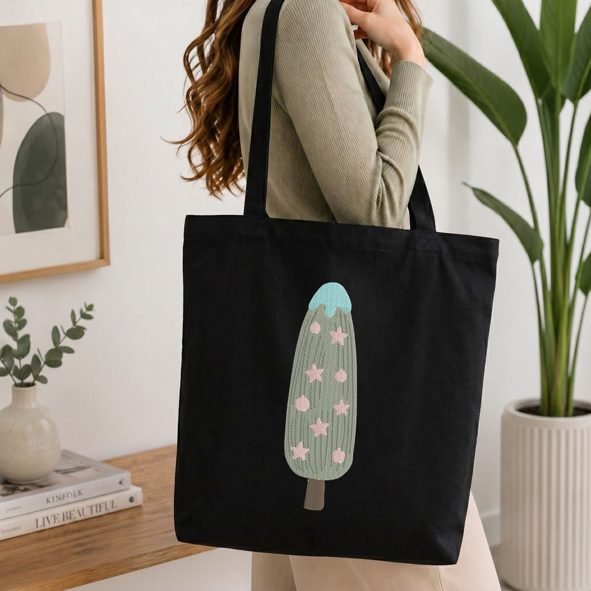 Woman carrying a black natural canvas tote bag featuring an embroidered Christmas tree design in a bright, modern interior setting.