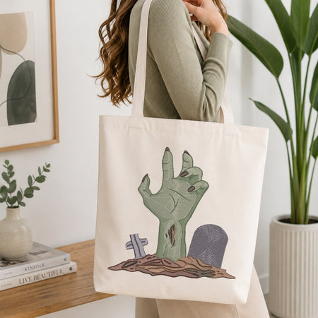 A woman wearing a sage green ribbed long-sleeve top carries a cream canvas tote bag with a zombie hand graphic on the front, set in a bright, modern room with a wooden table, books, and a potted plant.