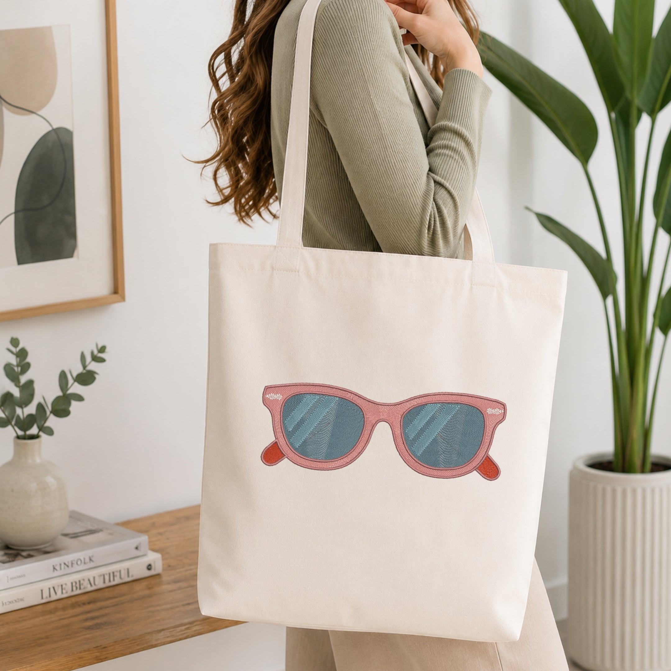 A woman wearing a sage green ribbed long-sleeve top carries a cream canvas tote bag with a pink sunglasses graphic on the front, set in a bright, modern room with a wooden table, framed art, and a potted plant.